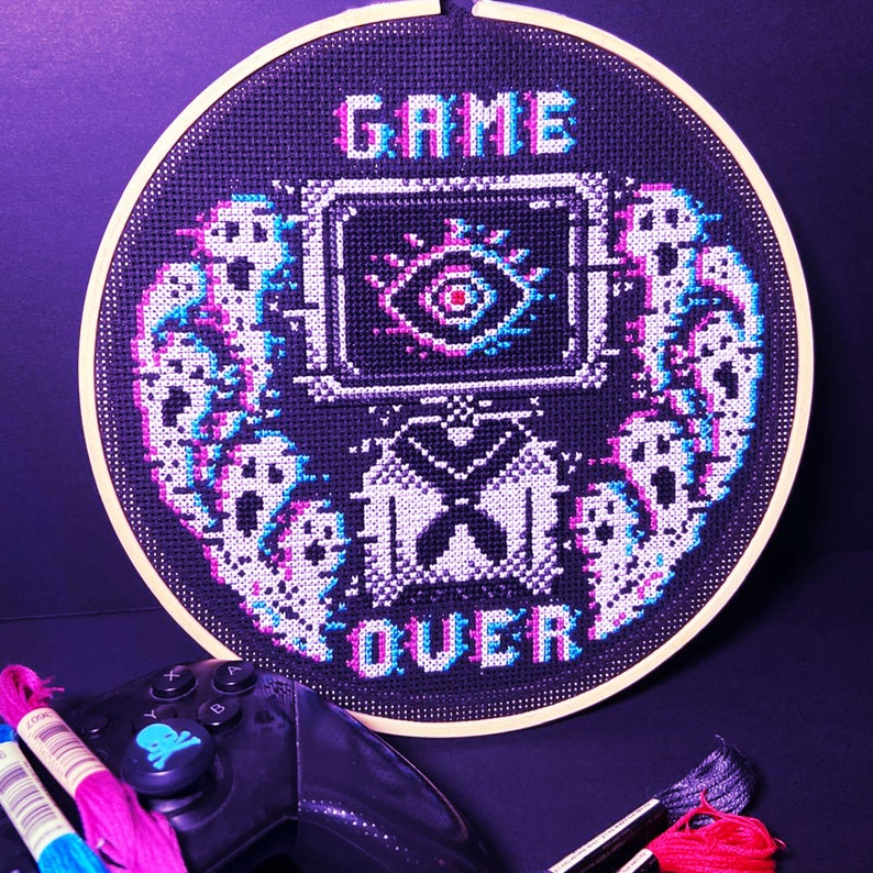 May include: A circular embroidery hoop with a black fabric background featuring a pixelated design of a game over screen. The design includes the words "GAME OVER" and a pixelated eye with a pink and blue color scheme.