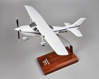 May include: A white Cessna 182 Skylane model airplane with a black propeller and dark blue trim. The fuselage displays the registration N182NU. It is mounted on a polished wooden base with a plaque reading Cessna 182 Skylane Scale 1:32.