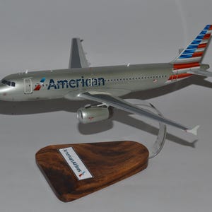 May include: A silver model airplane with the "American" logo on the side and a tail fin with red, white, and blue stripes. The model sits on a wooden base with a metal stand. The base has a label that reads "American Airlines".