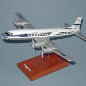 Può includere: Un modellino di aereo argentato e bianco con dettagli blu. L'aereo è un Douglas DC-7 e ha le parole "Pan American World Airways" sul lato. La coda dell'aereo ha le lettere "PAA" su di essa. L'aereo è seduto su una base di legno.