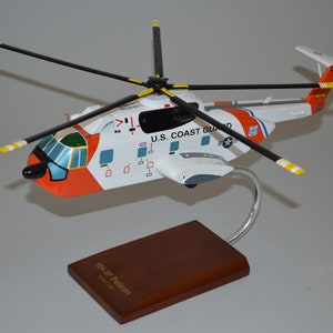Può includere: Un modello bianco, arancione e verde di un elicottero HH-65C Pelican della Guardia Costiera degli Stati Uniti con pale nere e un supporto argentato. L'elicottero ha le parole "U.S. COAST GUARD" stampate sul lato.