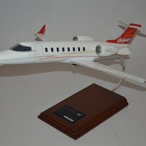 May include: A white Learjet 45 model airplane with red accents and the registration N45LJ on the side. The model is on a wooden base with a Learjet 45 label.