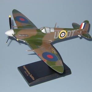 May include: A model of a Supermarine Spitfire airplane. The aircraft is painted in camouflage colors of green and brown, with roundels and markings. The model is mounted on a black base that reads "Supermarine Spitfire Scale 1/32".