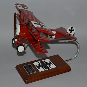 Pode incluir: Um modelo de avião vermelho e branco com uma cruz de ferro preta na cauda. O modelo está em uma base de madeira com um suporte de metal.