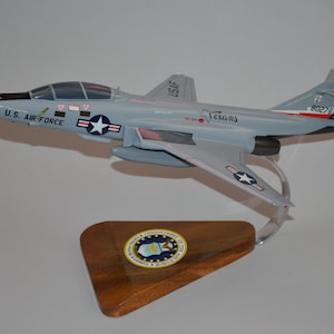 Può includere: Modello di aereo F-4 Phantom II grigio dell'US Air Force, con la scritta "Texans" e il numero "80276", esposto su un supporto di legno con un emblema dorato.