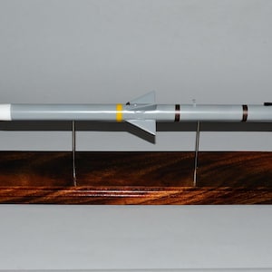 May include: A gray and white model of a missile on a wooden stand. The missile has a yellow stripe and two black stripes.