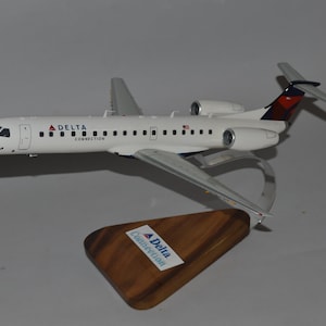 Può includere: Un modello di aereo Delta Connection bianco con ali e alettoni argentati. Il modello è montato su un supporto trasparente e una base in legno con il logo Delta Connection. La coda presenta dettagli rossi e blu.