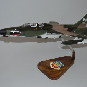 May include: A model of a green, brown, and white F-105 Thunderchief fighter jet with a shark mouth on the nose. The jet is on a wooden stand with a patch for the 332nd Fighter Squadron. The tail of the jet has the text "WW 320".