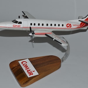 May include: A model airplane, white with red accents, featuring the Comair logo and tail markings. The plane is mounted on a wooden base with the Comair logo. The model has two propellers and is displayed on a silver stand.
