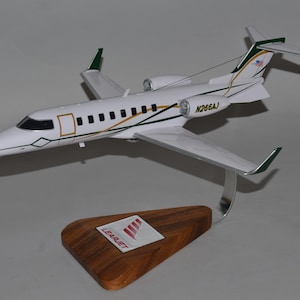 May include: A white and green Learjet model airplane with the registration number N266AJ. The model is mounted on a wooden base with the Learjet logo. The plane has a sleek design with detailed features.