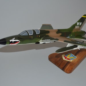 May include: A green, brown, and white model airplane with a shark tooth design on the nose. The plane is on a wooden stand with a military insignia. The tail of the plane has the letters "WW" and the number "320" on it.