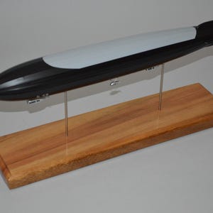 May include: A model of a zeppelin airship, black and white in color, mounted on a wooden base. The airship is supported by thin metal rods. The wooden base is rectangular and has a natural wood grain finish.