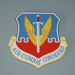 Può includere: Un emblema colorato con una spada e ali, con il testo "AIR COMBAT COMMAND". L'emblema ha uno sfondo blu, dettagli rossi e gialli e un bordo bianco. Il design ha la forma di uno scudo.