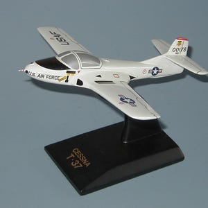 以下が含まれることがあります： 黒い台座に展示された、US Air Forceのマーキングが施された白いセスナT-37モデル飛行機。 このモデルは、詳細な翼、尾翼、コックピットを備え、ベースには「Cessna T-37」の文字が記されています。