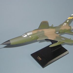May include: A model of an F-105D fighter jet, featuring a camouflage pattern of green and brown. The model is mounted on a black base, with the text "F-105D SCALE 1/48" visible. The jet has a silver cockpit and the number "6771" on the tail.