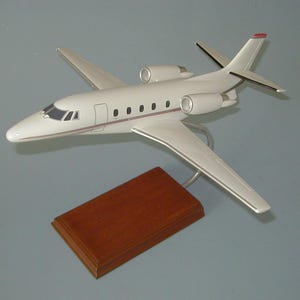May include: A white model airplane with a sleek fuselage, two rear-mounted jet engines, and a thin red stripe along the side. The aircraft is mounted on a silver metal stand attached to a rectangular, polished brown wooden base for display.