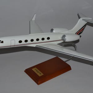 Könnte beinhalten: Ein weißes Modellflugzeug mit schwarzen Bullaugen, roten und schwarzen Akzenten und silbernen Flügeln. Das Modell ist auf einem Holzsockel mit einer goldenen Plakette montiert. Das Flugzeug ist eine Gulfstream G550.
