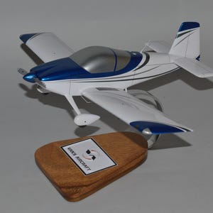 May include: A white and blue model airplane with a silver canopy, sitting on a wooden base with the text "Vans Aircraft".