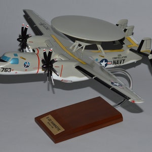 May include: A gray and white model airplane with black propellers and a rotating radar dish on top. The model is labeled "E-2D Hawkeye" and "Scale 1:48". It also has the text "USS Constellation Navy" on the side.