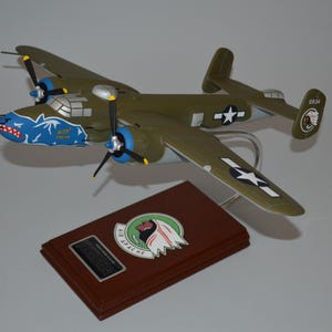 May include: A green and brown model aircraft with blue and white markings. The plane has a shark mouth painted on the nose and the text "82nd Airborne" on the side. The plane is on a wooden base with a plaque that reads "82nd Airborne".