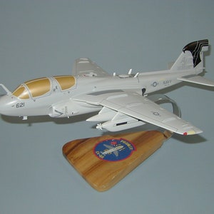 May include: A white model airplane with a black and white tail. The plane is on a wooden stand with a blue and white emblem that says "Prospector EA-6B".