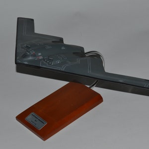 May include: A gray model of a Northrop Grumman B-2 Spirit stealth bomber on a wooden base. The model has a silver stand and the base has a label that reads "Northrop Grumman B-2 Spirit 1/72 Scale".