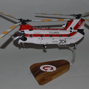 Op de afbeelding: Een modelhelikopter in rood en wit, met het woord "COLUMBIA" en het nummer "2CH". De rotorbladen hebben gele en witte strepen. Het staat op een houten voet met een logo.