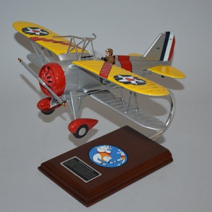 May include: A yellow and silver model airplane with red accents. The plane is on a wooden base with a round emblem featuring a cartoon character. The plane is a biplane with a single propeller.
