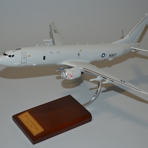 May include: A white model airplane with a navy blue "NAVY" inscription on the side. The model is on a wooden stand with a plaque that reads "Boeing P-8A Poseidon Scale 1:72".