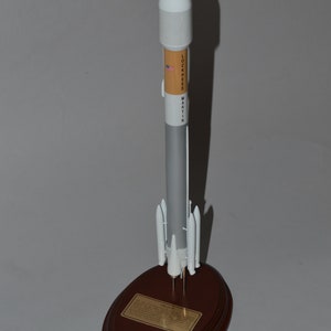May include: A model rocket with a white nose cone, a tan and gray body, and white boosters, mounted on a wooden base. The rocket has the text "LOCKHEED MARTIN" and a small American flag. A gold plaque is on the base.