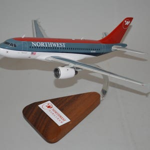 May include: A model airplane of a Northwest Airlines aircraft. The plane is primarily blue and white with a red tail and the Northwest logo. It is mounted on a wooden base with a metal support.