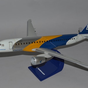 Può includere: Un modello di aereo Embraer E190-E2, con una livrea bianca, gialla e blu. Il modello è esposto su un supporto blu con il logo Embraer. L'aereo ha ali argentate e dettagli.