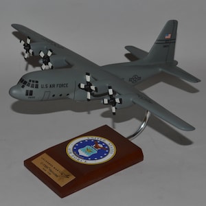 Könnte beinhalten: Ein graues C-130 Hercules Modellflugzeug der U.S. Air Force mit vier Propellern, montiert auf einem silbernen, geschwungenen Ständer auf einem rechteckigen Holzsockel mit kreisförmigem Air Force-Emblem und einer kleinen goldenen Plakette.