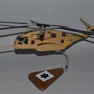 May include: A tan and black model helicopter with a black star on the tail. The helicopter is on a wooden stand with a black star on it. The helicopter is a Sikorsky CH-53 Sea Stallion.