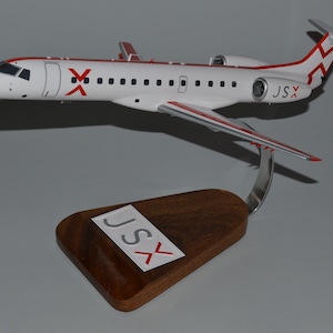 Può includere: Un modello di aereo bianco e rosso con la scritta "JSX" sulla coda e sulla base. Il modello è montato su una base di legno con un supporto metallico. L'aereo ha dettagli rossi e un design a "X" rosso sulla fusoliera.
