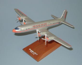 Könnte beinhalten: Ein silbernes und orangefarbenes Flugzeugmodell eines Douglas DC-7, mit der Aufschrift "AMERICAN AIRLINES" an der Seite, auf einem Holzsockel. Der Sockel trägt die Aufschrift "Douglas DC-7 Maßstab 1/100".