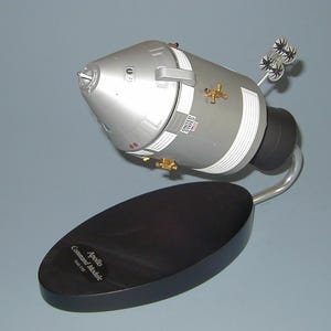 May include: A silver and white model of an Apollo Command Module on a black stand. The model has a United States flag on the side and the text "Apollo Command Module Scale 1:144" on the stand.