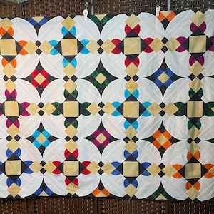 May include: A quilt with a repeating pattern of white, black, orange, red, green, blue, and yellow squares and circles. The quilt is made of fabric and has a quilted texture.
