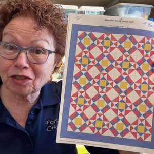 May include: A woman holds up a printed pattern for a quilt. The pattern is called "GOV Ohio Star Design Quilt" and has a blue background with red, yellow, and white squares and triangles. The pattern is for a quilt with a 4 inch border.