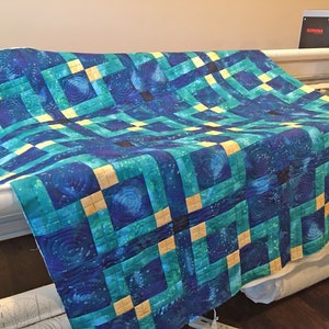 Puede incluir: Una colcha azul y verde con un patrón geométrico de cuadrados y espirales. La colcha se está acolchando en una máquina de acolchar de brazo largo.