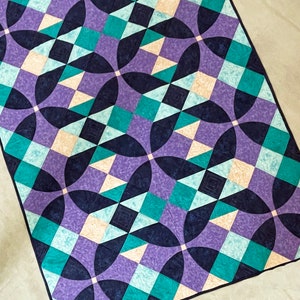 Puede incluir: Una colcha con un patrón geométrico en tonos de morado, verde azulado y crema. La colcha está hecha de cuadrados y círculos, creando un diseño visualmente interesante.
