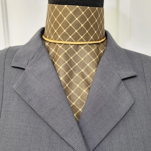 Pode incluir: Um blazer cinza-carvão com um padrão de grade sutil usado sobre um ascot marrom com uma estampa de diamantes e pontos na cor creme. Um cordão fino de tom dourado metálico está amarrado ao redor do pescoço do ascot, sobre o tecido.