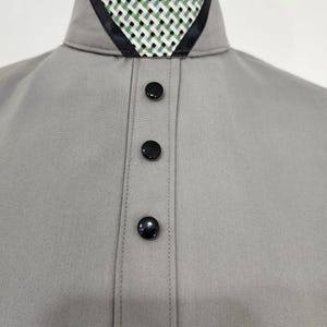 Pode incluir: Uma camisa cinza-claro com gola alta e um inserto de padrão tecido em preto e branco. A carcela frontal é adornada com três botões redondos de cor preta brilhante. O tecido tem uma textura lisa e sólida, com costuras bem definidas.