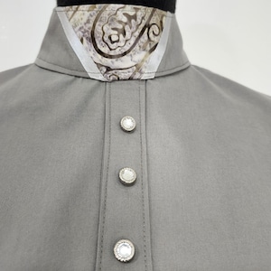 Puede incluir: Una camisa gris sólida con cuello alto y un inserto con estampado de cachemira en blanco y gris. La tapeta frontal está adornada con tres botones circulares de tono plateado, cada uno con un cristal transparente facetado en el centro.