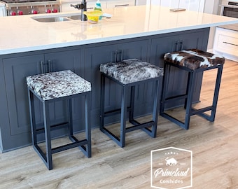 Puede incluir: Cuatro taburetes de bar cuadrados con marcos de metal negro y asientos tapizados con varios patrones de piel de vaca en tonos blanco, marrón, gris y negro. Los taburetes están colocados a lo largo de una isla de cocina gris oscuro.