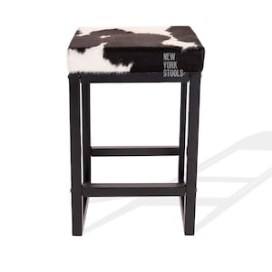 Puede incluir: Un taburete cuadrado con asiento de piel de vaca en blanco y negro y estructura de metal negro. El asiento presenta el texto NEW YORK STOOLS impreso en blanco. El taburete tiene cuatro patas rectas unidas por barras de soporte horizontales.
