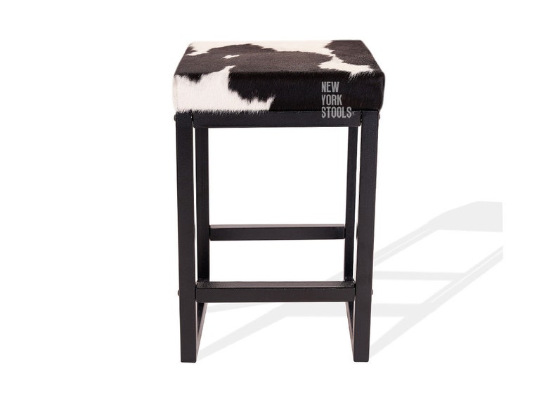 Puede incluir: Un taburete cuadrado con asiento de piel de vaca en blanco y negro y estructura de metal negro. El asiento presenta el texto NEW YORK STOOLS impreso en blanco. El taburete tiene cuatro patas rectas unidas por barras de soporte horizontales.