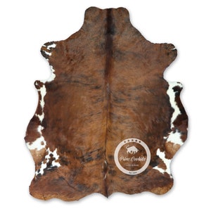 Puede incluir: Alfombra de piel de vaca natural con un patrón marrón y blanco. La alfombra tiene una forma irregular y una textura visible, con una mezcla de tonos marrones oscuros y claros. El logotipo "Prime Cowhides" está presente.