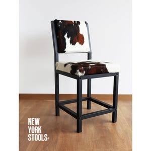 Könnte beinhalten: Ein schwarzer Barhocker aus Metall mit einer Sitzfläche und Rückenlehne aus weißem und braunem Kuhfell. Der Hocker hat eine quadratische Basis und ein schlichtes Design. "NEW YORK STOOLS" ist auf dem Boden aufgedruckt.