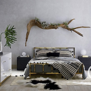 K&ouml;nnte beinhalten: Ein Bettgestell aus goldenem Metall mit einer schwarz-wei&szlig;en gestreiften Bettdecke und einer &Uuml;berwurfdecke. Das Bett steht in einem Schlafzimmer mit einer wei&szlig;en Kommode, einem Treibholzast mit Pflanzen und einem runden Spiegel.